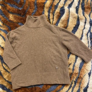 Beige polotröja från Emma och Malena - Mysig beige polotröja från Emma och Malena i mjuk ullmix. Tröjan har trekvartsärmar och en hög polokrage. Liten broderad ankardetalj nertill ger en stilren touch. Perfekt för lager-på-lager under kyliga dagar.                          Tröjan har vissa skavanker, i form av pyttersmå slitagehål(bild 3,4 på framsidan & 6,7 på baksidan av tröjan) 