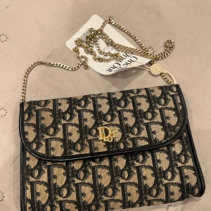 Dior axelväska med kedja och monogram - Snygg axelväska från Dior med klassiskt monogrammönster i svart och beige. Väskan har guldfärgad kedja som axelrem och ett guldfärgat Dior-emblem på framsidan. Materialet är textil med detaljer i skinn och väskan stängs med tryckknapp.