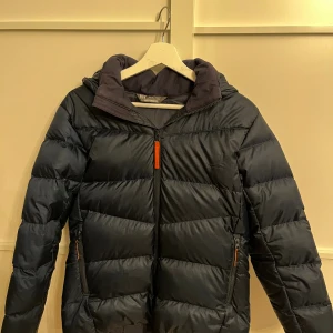 Marinblå pufferjacka från Helly Hansen - Säljer en marinblå pufferjacka från Helly Hansen med huva och orange detaljer på dragkedjorna. Jackan är vadderad och har en sportig look, perfekt för kalla dagar. Klassisk passform och hel dragkedja framtill. Odinare pris 3000kr, nämn ditt pris är öppen för allt🙌🏼