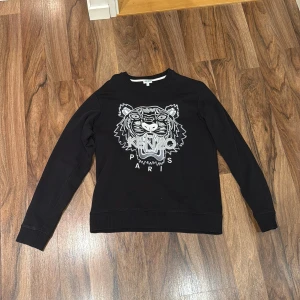 Svart Kenzo sweatshirt med tigerbrodyr - Svart sweatshirt från Kenzo med ikonisk tigerbrodyr och Kenzo Paris-text framtill. Tröjan har rund hals, långa ärmar och ribbade muddar. Perfekt statement-plagg för dig som vill sticka ut med en snygg och trendig look.