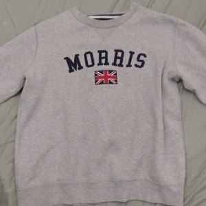 Grå sweatshirt från Morris med tryck - Grå sweatshirt från Morris med mörkblå text och brittisk flagga på bröstet. Klassisk rund halsringning, ribbade muddar och en liten patch på ärmen. Skön och stilren tröja i bomull, perfekt för en avslappnad look.