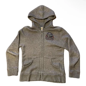 Grå hoodie från Hysteric Glamour - Grå hoodie från Hysteric Glamour med dragkedja, huva och stora tryck både fram och bak. Tröjan har ribbade muddar och fickor framtill. Tillverkad i 100% bomull och har en avslappnad passform. Perfekt för dig som gillar streetwear vibes.