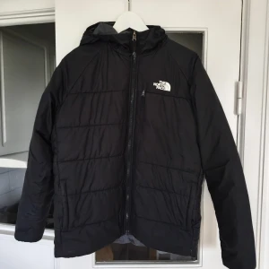 Svart pufferjacka från The North Face - Svart pufferjacka från The North Face med huva och dragkedja framtill. Jackan har en enkel, stilren design med logga på bröstet och ryggen. Perfekt för kyliga dagar och enkel att matcha med olika outfits.