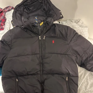 Svart pufferjacka Polo Ralph Lauren M - Svart pufferjacka från Polo Ralph Lauren med röd broderad logga på bröstet. Jackan har huva med snörning, flera dragkedjeförsedda fickor och quiltad design. Perfekt för kalla dagar och riktigt snygg streetstil.