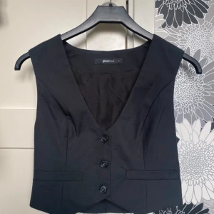 Svart klassisk väst från Gina Tricot - Snygg svart väst från Gina Tricot i klassisk modell med djup v-ringning och tre knappar framtill. Västen har en figurnära passform och är tillverkad i ett slätt polyestertyg. Perfekt att styla över en skjorta eller t-shirt för en trendig look.