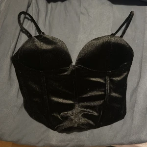 Svart sammets korsett från Gina Tricot - Säljer en svart korsett från Gina Tricot i storlek XS. Toppen är gjord i mjuk sammet med formade kupor, justerbara axelband och dragkedja bak. Snygg croppad passform som ger en cool siluett. Perfekt för att lyfta din outfit! Testad men aldrig använd. 🖤