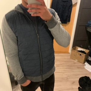 Unik cardigan jacka med dragkedja - Säljer en stilren pufferjacka med grå ärmar och blå kropp. Jackan har hel dragkedja framtill, ribbade muddar och huva. Perfekt för dig som vill ha en chill och enkel look till vardags. Passar dig som gillar bekväma och snygga jackor. Nypris:1399Kr
