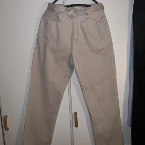 Beiga byxor med knyt från NLY Trend - Snygga beiga byxor från NLY Trend i storlek 36. Byxorna har hög midja, raka ben och ett bälte med knyt i midjan. Tillverkade i bomullstyg med fickor fram och bak. Perfekta för en chill och stilren look.