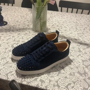 Mörkblå sneakers - Säljer ett par mörkblå sneakers från Christian Louboutin i lyxig mocka med coola nitar på tån och klassisk röd sula. Skorna har snörning och vit platt sula, insida i beige läder. Perfekt för dig som vill sticka ut med exklusiv design.
