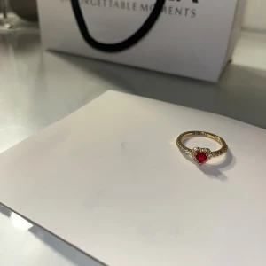 Pandora ring med rött hjärta - Superfin guldfärgad ring från Pandora med ett rött hjärta i mitten och små gnistrande stenar längs bandet. Säljer den för den va för stor för mig använt bara två dagar. Pandora box och påse följer även med. Har tyvär inte kvitto eftersom jag beställde. Därför säljer jag bara för 499kr