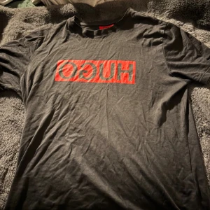 Svart HUGO t-shirt med röd logga - Cool svart t-shirt från HUGO med stor röd logga tryckt på bröstet. Klassisk passform och rund halsringning, perfekt för en avslappnad streetstyle-look. Tillverkad i mjuk bomull för skön känsla hela dagen.