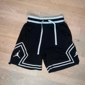 Svarta Jordan shorts med vita detaljer - Snygga svarta Jordan shorts med vita ränder och logga på benen. De har elastisk midja med vit snörning och är tillverkade i ventilerande polyester mesh. Perfekta för sport eller chill, med loose passform och cool design.