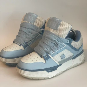 AMIRI ljusblå och vita sneakers - Säljer ett par snygga AMIRI sneakers i ljusblått och vitt med meshdetaljer och tydlig logga på sidan och hälen. Skorna har snörning, platt sula och rund tå. Perfekt för dig som vill sticka ut med stilrena och trendiga sneakers.