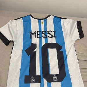 Argentina Messi fotbollströja Adidas S - Säljer en officiell Argentina fotbollströja från Adidas i storlek S, med blå och vita vertikala ränder, svarta detaljer och tryck av Messi nummer 10 på ryggen. Framsidan har FIFA 2022-märke, AFA-logga och motiv av Messi med VM-pokalen. Tillverkad i lätt och ventilerande Climalite-material.