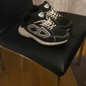 Svarta Dior sneakers med vita detaljer - Säljer ett par svarta Dior sneakers med vita och silverfärgade detaljer samt tydlig CD-logga på sidan. Skorna har vit sula, snörning och en sportig, chunky design som sticker ut. Perfekta för dig som vill ha en lyxig och trendig look.Använd bara en gång 