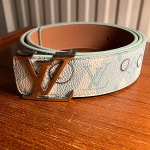 Ljusblå skinnbälte - Snyggt bälte i ljusblått och beige skinn med klassiskt LU-monogram och silverfärgat LU-spänne. Bältet har en bred form och exklusiv känsla, perfekt för att lyfta din outfit med en lyxig touch. Tillverkat i Frankrike med detaljerad präglad insida. Helt nytt!!