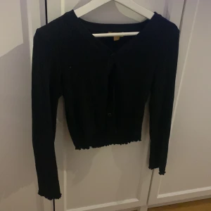 Svart ribbad kofta från Gina Tricot - Svart ribbad kofta från Gina Tricot med v-ringning och knappar framtill. Modellen är croppad med vågiga kanter vid ärmslut och nederkant. Perfekt för lager-på-lager och enkel att matcha med allt.