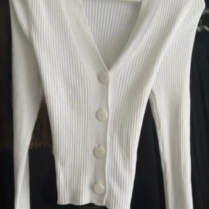 Vit ribbad tröja med knappar - Säljer en vit ribbad tröja med v-ringning och stora knappar framtill. Den funkar också som att använda som en kofta då den är lite lösare i midjan på mig. Köpt utomlands och har aldrig använt den nångång. 