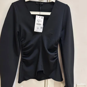  Ny Svart långärmad topp från Zara ny kollektion  - Stilren svart topp från Zara med v-ringning och rynkade detaljer längs sidorna framtill. Toppen har lång ärm och figurnära passform som framhäver siluetten. Perfekt att matcha med både jeans och kjol.