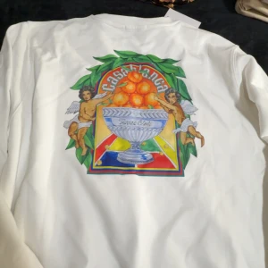 Vit sweatshirt från Casablanca - Vit sweatshirt från Casablanca med färgglad grafik på ryggen och mindre tryck fram. Motivet har änglar, apelsiner och en pokal med texten 'Tennis Club'. Tröjan har rund hals och är tillverkad i mjuk bomull. Perfekt för dig som gillar unika prints och streetstyle. Oanvänd, passar XL med. 