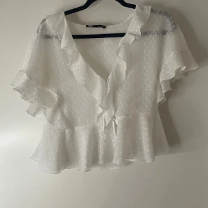 Vit blus med volanger från Zara - Säljer en söt vit blus från Zara i storlek S. Blusen har volanger längs v-ringningen och ärmarna, samt ett diskret prickigt mönster i tyget. Den är lätt och luftig med korta ärmar och omlottkänsla framtill. Perfekt för vår och sommar!