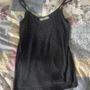 Svart linne från Dolce & Gabbana - Snyggt svart linne från Dolce & Gabbana med smala axelband dekorerade med strassdetaljer. Framsidan har små silvriga nitar som ger en cool touch. Mjukt material och figurnära passform, perfekt till jeans eller kjol. Ät liten i storleken!! Många pärlor som har ramlat av !!