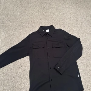 Svart overshirt från Selected Homme - Stilren svart overshirt från Selected Homme med klassisk krage, två bröstfickor med knappar framtill. Perfekt lager-på-lager-plagg med lång ärm och snygg, enkel design. Nypris 800kr 