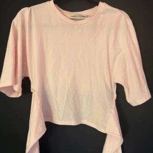 Ljusrosa croppad topp med knyt - Säljer en ljusrosa croppad topp i mjuk bomull med korta ärmar och snygg knytdetalj framtill. Perfekt för dig som gillar en avslappnad och trendig stil. Enkel att matcha med både jeans och kjol.