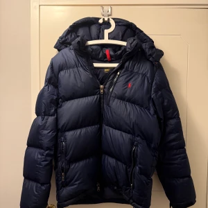 Mörkblå pufferjacka från Polo Ralph Lauren - Säljer en mörkblå pufferjacka från Polo Ralph Lauren med huva och dragkedja. Jackan är väldigt varm och skön nu till vinter men säljer den då jag köpt en ny. Nypris cirka 6000