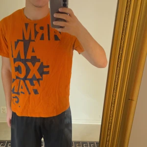Orange t-shirt från Armani Exchange XL - Snygg orange t-shirt från Armani Exchange i storlek XL som har krympt till en L/M/S (kan töjas ut).T-shirten har ett stort svart grafiskt tryck med texten 'ARMANI EXCHANGE' på både fram- och baksidan. Klassisk passform med rund hals och korta ärmar. Perfekt för dig som vill sticka ut med färg och stil. Pris kan diskuteras