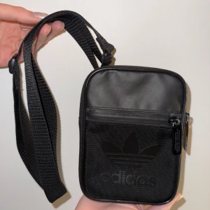 Svart axelväska från Adidas - Svart axelväska från Adidas med justerbar rem och dragkedja. Väskan har Adidas logga framtill, meshdetalj på framsidan och textdetalj på baksidan. Tillverkad i slitstark polyester och har ett huvudfack samt ytterfack med dragkedja.
