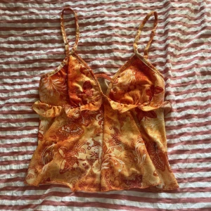 Orange blommig linnetopp med volang - Säljer en snygg orange linnetopp med blommigt mönster och tunna axelband. Toppen har volangdetaljer under bysten och är tillverkad i ett lätt, transparent syntetmaterial. Perfekt för dig som gillar färgstarka och unika plagg med sommarkänsla!