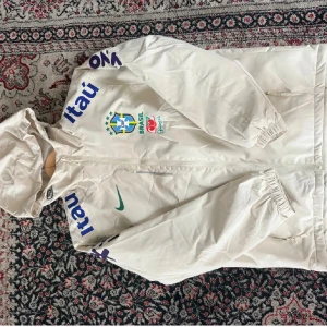 Vit Nike Brasil jacka med huva - Vit jacka från Nike med Brasil-logga och sponsormärken. Jackan har huva, dragkedja framtill och elastiska muddar. Snygga blå detaljer på ärmarna och grönt Nike-märke på bröstet. Perfekt för dig som gillar fotboll och streetstyle. Väldigt varm på insidan. Står att den är storlek M men den sitter som S och passar också dig som har XS .Den är i fint skick och oanvänd, tveka inte att höras av vid intresse😊 ( den sitter mer som än jacka än windbreaker )