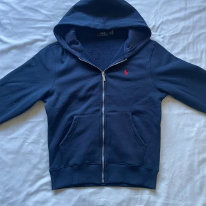 Ralph Lauren zipup mörkblå - Marinblå hoodie från Polo Ralph Lauren med dragkedja och klassisk röd broderad logga på bröstet. Tröjan har huva, känguruficka och är tillverkad i mjuk bomull. Perfekt för en chill och stilren look.
