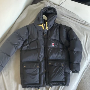 Svart dunjacka från Fjällräven M - Säljer en svart dunjacka från Fjällräven i storlek M. Ganska tjock är den så jättevarm o gosig, stora fickor med tryckknappar och klassisk Fjällräven-logga på bröstet.  Storlek M (säkert L också) i dam passar den, kan passa S också men den kan den se lite stor ut, den är i väldigt fint skick nästan nyskick men saknar tyvärr 2 av 4 vita grejerna på snörena.     Expedition Down lite nypris ≈7000kr o den är nästan i nyskick så nästan 50%/3000kr rabatt