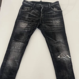 Svarta Dsquared2 jeans med slitningar - Säljer ett par svarta jeans från Dsquared2 med coola slitningar och trasiga detaljer på benen. Jeansen har en smal passform och klassisk femficksdesign. Materialet är jeans och färgen är tvättad svart med grå inslag för en snygg, använd look.