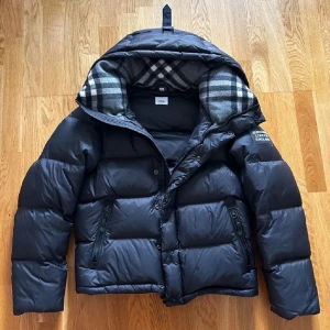 Burberry pufferjacka - Kvitto finns! Strl S men passar M också! Bara testad! Snygg svart pufferjacka/väst från Burberry med avtagbara ärmar och huva. Insidan av huvan har klassiskt rutigt Burberry-mönster i grått och vitt. Västen har två dragkedjefickor och knappar framtill. Perfekt för lager-på-lager och streetstyle.