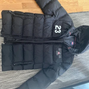 Svart dunjacka med huva - Svart dunjacka med huva och dragkedja. Jackan har broderad logga och siffran 23 på bröstet, samt två fickor med dragkedja. Klassisk pufferstil som håller dig varm under kalla dagar.
