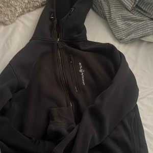 Svart hoodie från Sail Racing M - Svart hoodie från Sail Racing med dragkedja snett över bröstet och logga på vänster sida. Tröjan har huva, lång ärm och en bröstficka med dragkedja. Perfekt för dig som gillar sportig och clean stil.