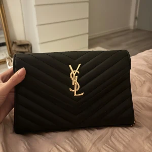 Svart quiltad axelväska från YSL - Säljer en svart quiltad axelväska från YSL med ikoniskt guldigt YSL-emblem framtill. Väskan har kedjerem i guld och elegant chevronmönster. Perfekt storlek för det viktigaste och ger en lyxig vibe till din outfit. Den är inte äkta!
