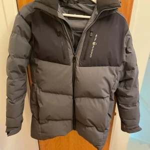 Grå pufferjacka från Sail Racing - Säljer en grå och mörkgrå pufferjacka från Sail Racing med huva och två dragkedjeförsedda bröstfickor. Jackan har en stilren look med logga på bröstet och ryggen, samt vadderad design som håller dig varm under kalla dagar.