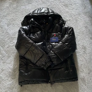 Ralph lauren pufferjacka  - Snygg svart pufferjacka från Polo Ralph Lauren med glansig finish och huva. Jackan har dragkedja framtill, två sidofickor med dragkedja och broderad logga på bröstet. Perfekt för kalla dagar och riktigt skön att bära. Den har även spännband så man kan justera storleken.