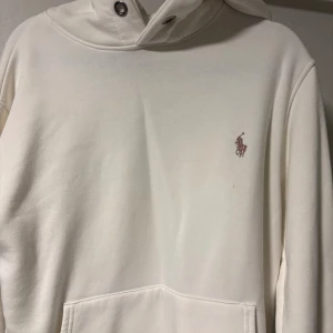 Vit hoodie från Ralph Lauren - Säljer en stilren vit hoodie från Ralph Lauren med broderad logga på bröstet. Tröjan har huva med metallöljetter och en klassisk känguruficka framtill. Perfekt för dig som gillar enkel och clean streetstyle.