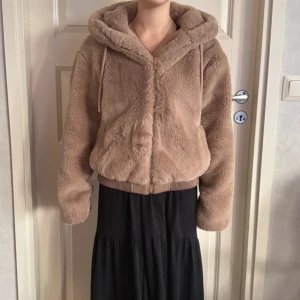 Brun teddyjacka med huva från Zara - Supermjuk teddyjacka från Zara i brun med huva och dragsko. Jackan har dragkedja framtill, ribbad mudd i midjan och långa ärmar. Perfekt för kalla dagar när du vill vara både varm och snygg.