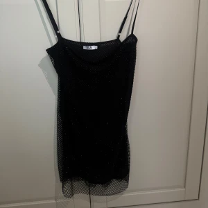 Svart glittrig meshklänning SLA The Label - Svart kort klänning från SLA The Label med glittrig mesh över hela och tunna axelband. Klänningen har ett nätmönster och sitter snyggt med en lätt draperad halsringning. Perfekt för dig som vill sticka ut med en cool och trendig look.