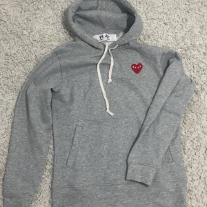 Grå hoodie Comme des Garçons Play - Grå hoodie från Comme des Garçons Play med klassiskt rött hjärta på bröstet. Tröjan har huva med vit snörning, känguruficka och långa ärmar. Mjuk bomullskvalitet och stilren design som passar perfekt till streetwear.