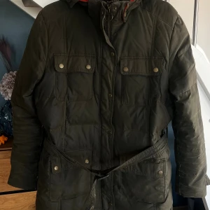 Mörkgrön dunjacka från Barbour - Snygg mörkgrön wax dunjacka från Barbour International med bälte i midjan, fyra fickor framtill med tryckknappar och huva. Jackan är vadderad med naturligt dun och fjädrar, perfekt för kalla dagar. Klassisk Barbour-logga på ärmen och dragkedja framtill.