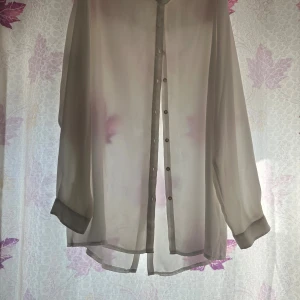 Beige transparent skjorta med knappar - Säljer en beige, transparent skjorta med långa ärmar och knappar framtill. Skjortan har en loose passform och är tillverkad i ett tunt, lätt material som ger en luftig känsla. Perfekt för lager-på-lager-stil.