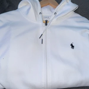 Vit hoodie från Polo Ralph Lauren - Vit hoodie från Polo Ralph Lauren med dragkedja framtill, huva med snörning och klassisk broderad logga på bröstet. Mjuk bomullskänsla och känguruficka framtill. Perfekt för en clean och stilren look.