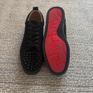 Christian Louboutin svarta sneakers med nitar - Svarta sneakers från Christian Louboutin i mocka med ikonisk röd sula och svarta nitar på tån. Skorna har snörning och rund tå, samt lyxig känsla med läderdetaljer invändigt. Perfekta för dig som vill sticka ut med edgy och exklusiv stil.